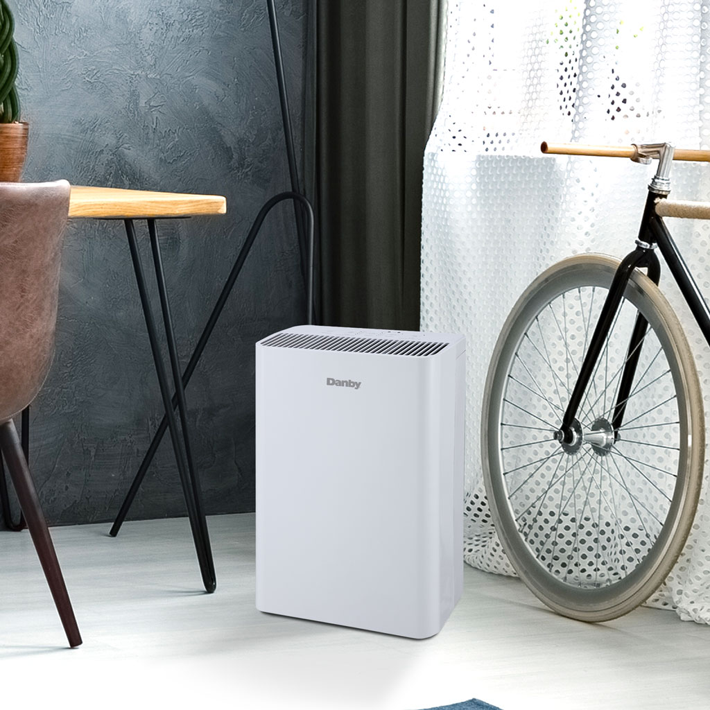Danby appliances air purifier 210 sq ft DAP152BAW-I shown in a home interior setting beside a table and bicycle, highlighting compact design for small living spaces