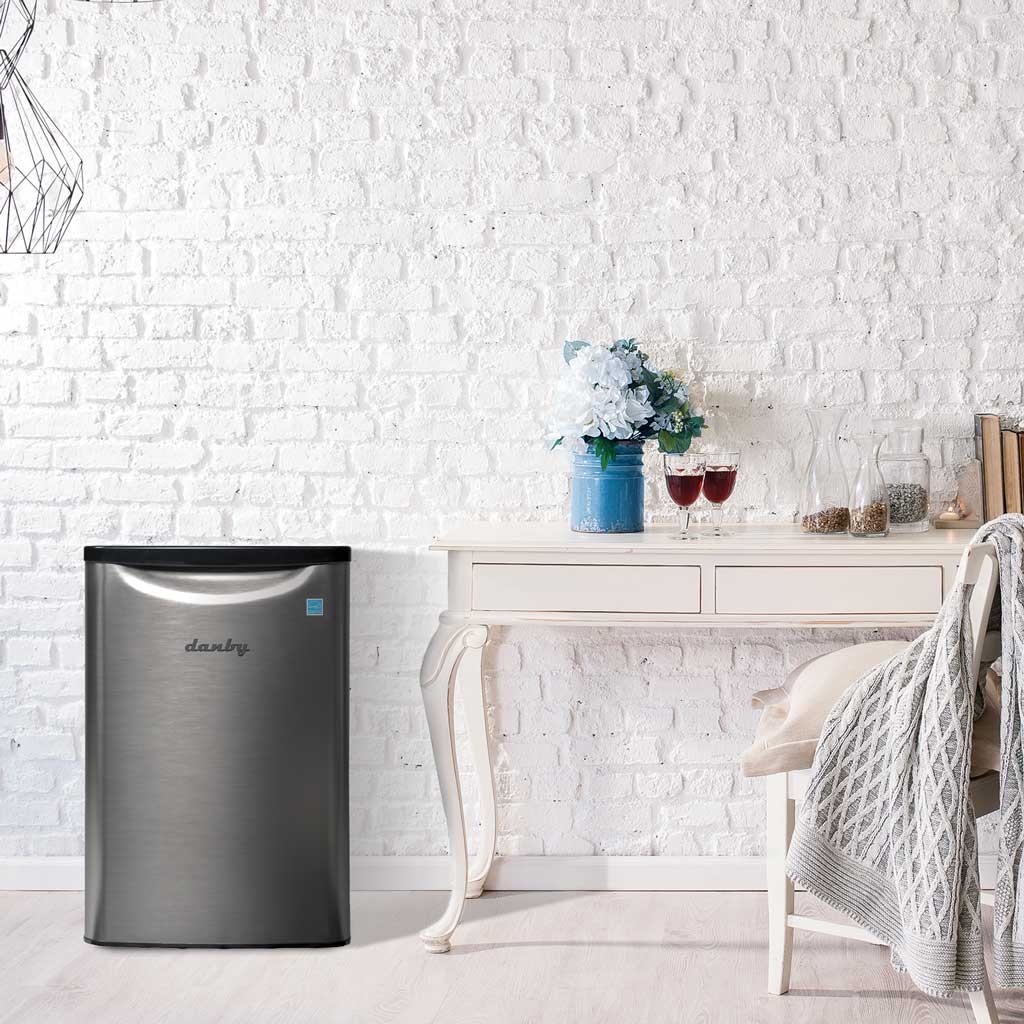 Danby appliances 2.6 cu ft compact fridge stainless DAR026A2BSLDB shown in a home interior beside a table and chair with glassware and décor, highlighting compact placement and everyday use