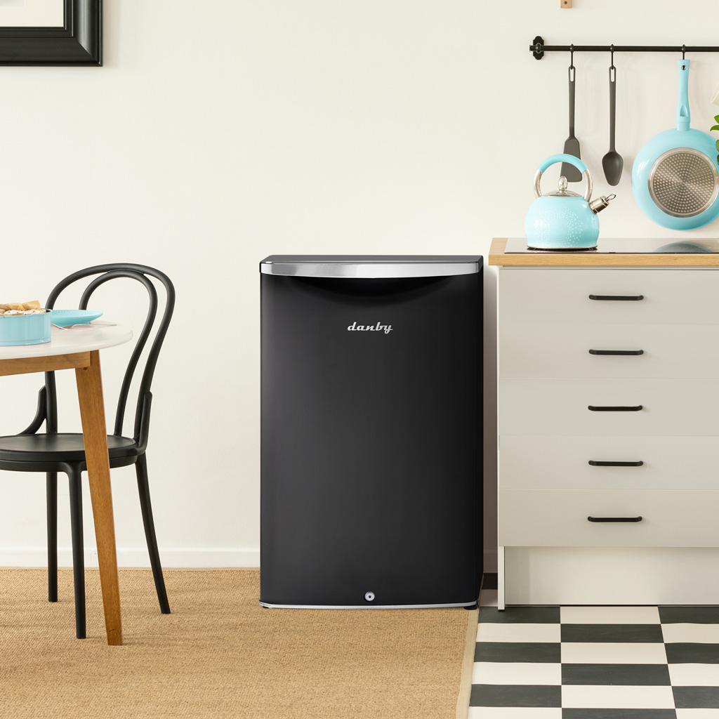 Danby appliances 4.4 cu ft compact fridge black DAR044A6MDB shown under a kitchen counter beside cabinets and wall décor highlighting compact placement and everyday use