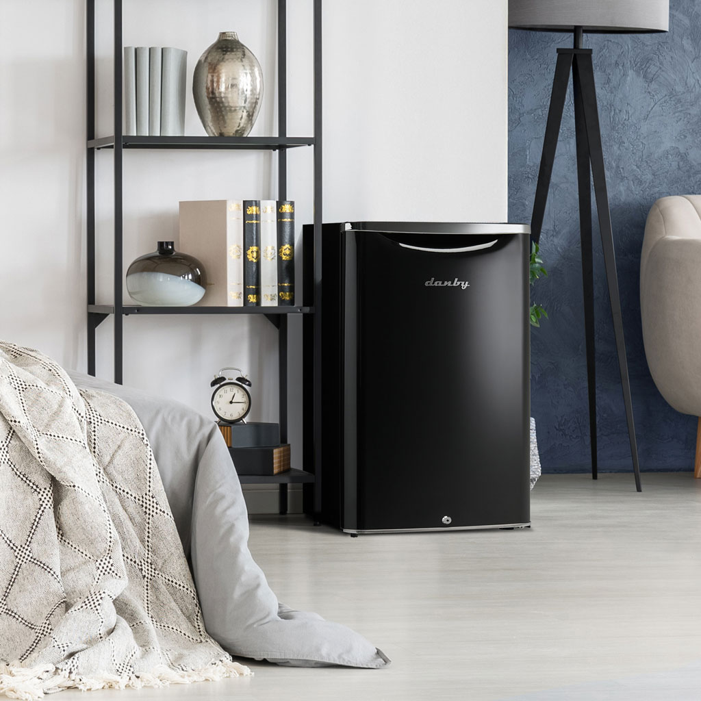 Danby appliances 4.4 cu ft compact fridge black DAR044A6MDB shown beside shelving and bedding in a home setting highlighting compact placement and modern design