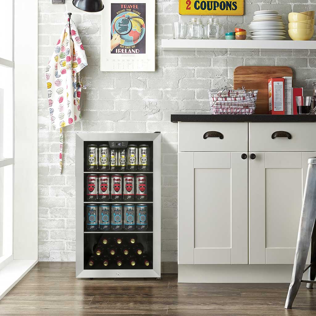 Danby appliances 4.5 cu ft beverage center stainless DBC045L1SS shown in a kitchen setting beside cabinetry and wall décor, displaying interior shelves stocked with beverage cans and bottles