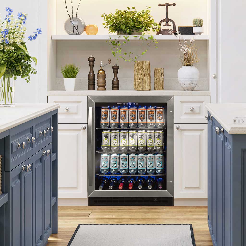Danby appliances 5.7 cu ft built in beverage center stainless DBC057A1BSS installed under a kitchen counter with blue cabinetry and wood countertop, showing interior shelves stocked with beverage cans