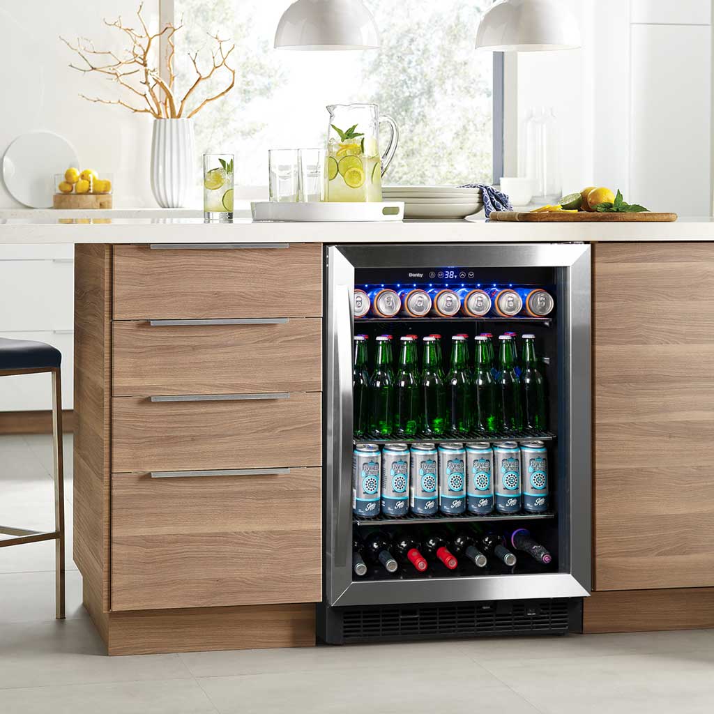 Danby appliances 5.7 cu ft built in beverage center stainless DBC057A1BSS installed under a kitchen counter with blue cabinetry and wood countertop, showing interior shelves stocked with beverage cans