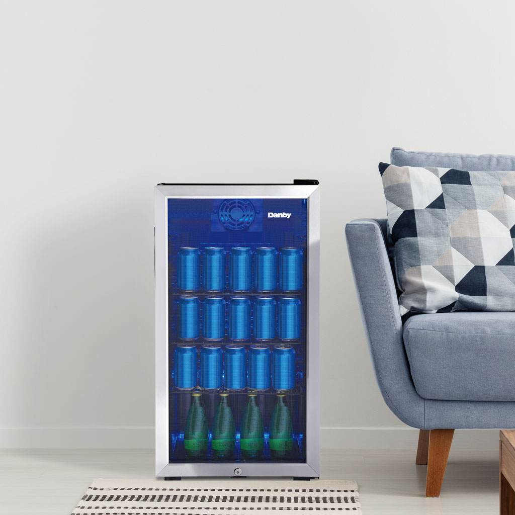 Danby appliances 3.1 cu ft beverage center stainless DBC117A1BSSDB-6 shown in a living room beside a sofa and wall vent, displaying interior shelves stocked with beverage cans and bottles