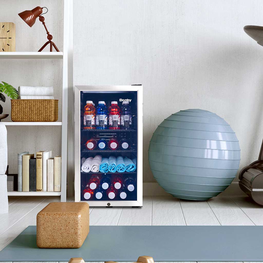 Danby appliances 3.1 cu ft beverage center stainless DBC117A1BSSDB-6 shown in a home setting beside shelving and decorative items, displaying interior shelves stocked with beverage cans and bottles
