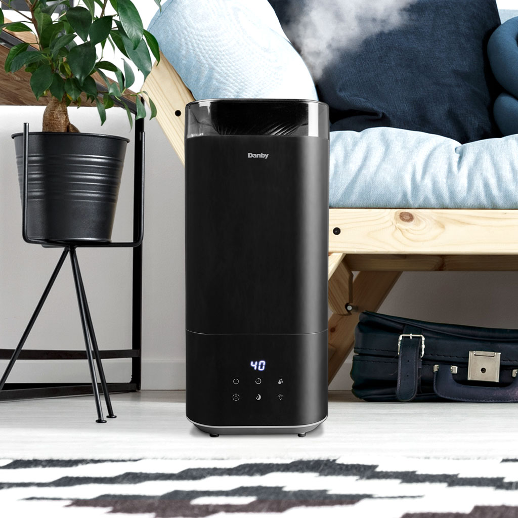 Danby appliances 5L ultrasonic top fill humidifier black DBHR13211BDD1 shown beside a bed and plant stand releasing visible mist in a bedroom setting on a patterned rug