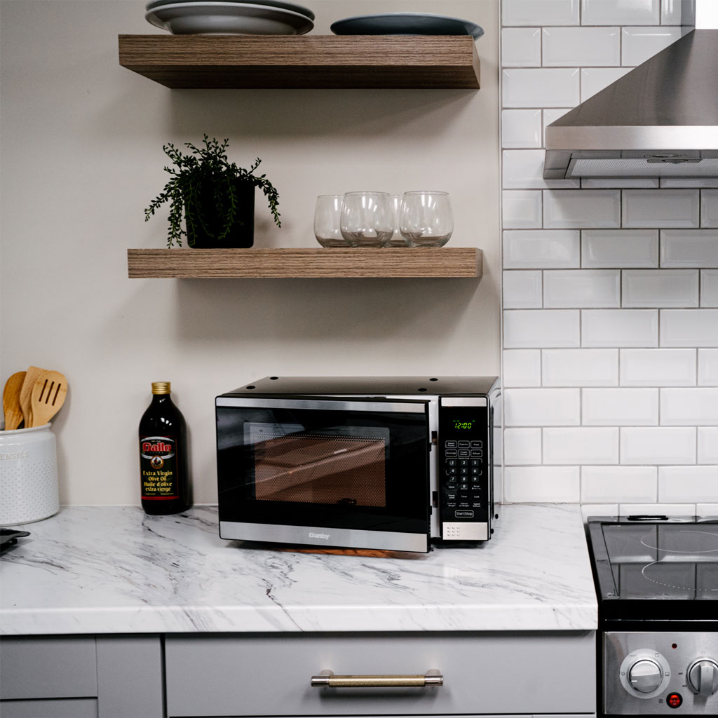 Danby appliances 0.7 cu ft under cabinet microwave DDMW007501G1 placed on a kitchen countertop with open wood shelving, accessories, and white tile backsplash