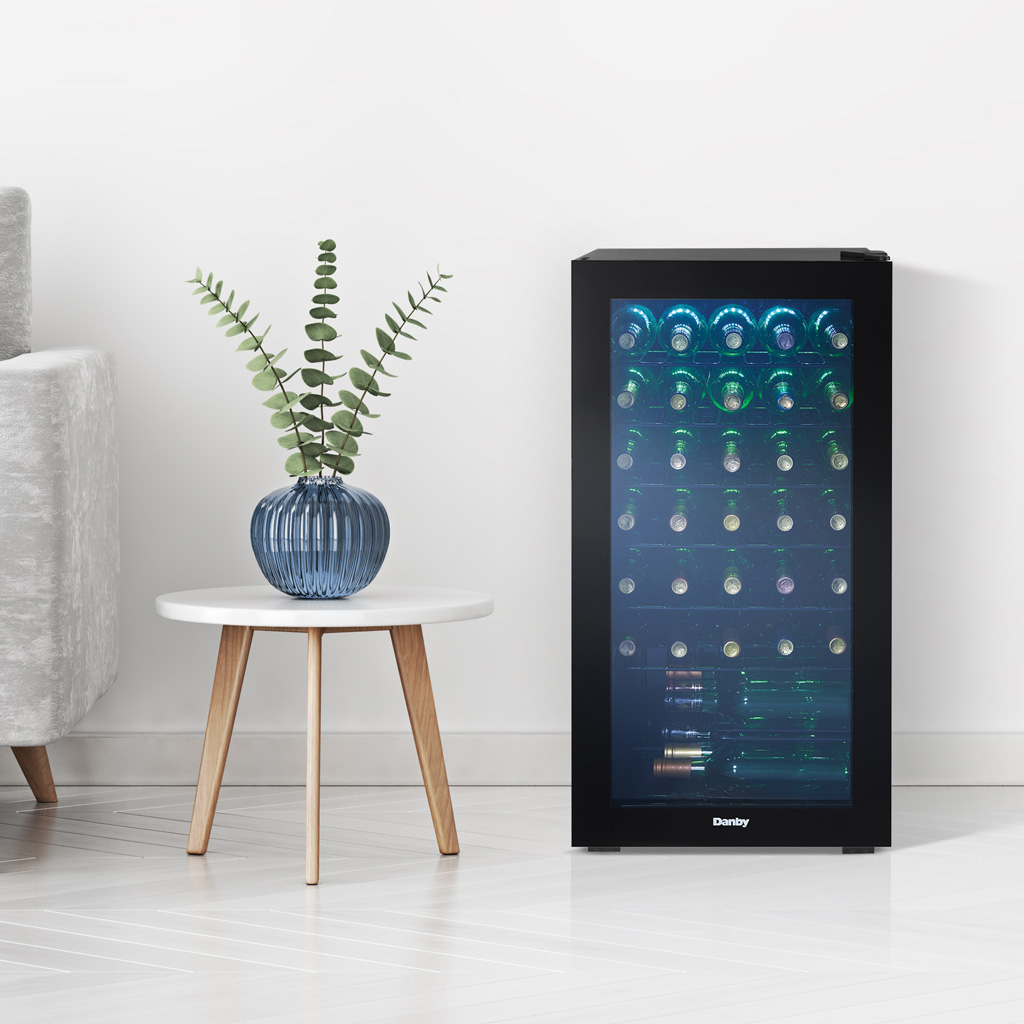 Danby 36 Bottle Free-Standing Wine Cooler in Black 3 Danby appliances 36 bottle wine cooler black DWC036A2BDB-6 shown in a minimalist room beside a stool and plant, displaying interior shelves with wine bottles visible through glass door