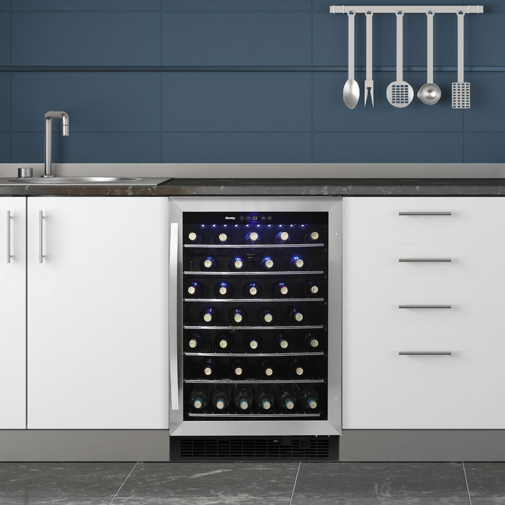 Danby appliances 60 bottle built in wine cooler stainless DWC057A1BSS installed under a kitchen counter with blue cabinetry, showing interior shelves stocked with wine bottles